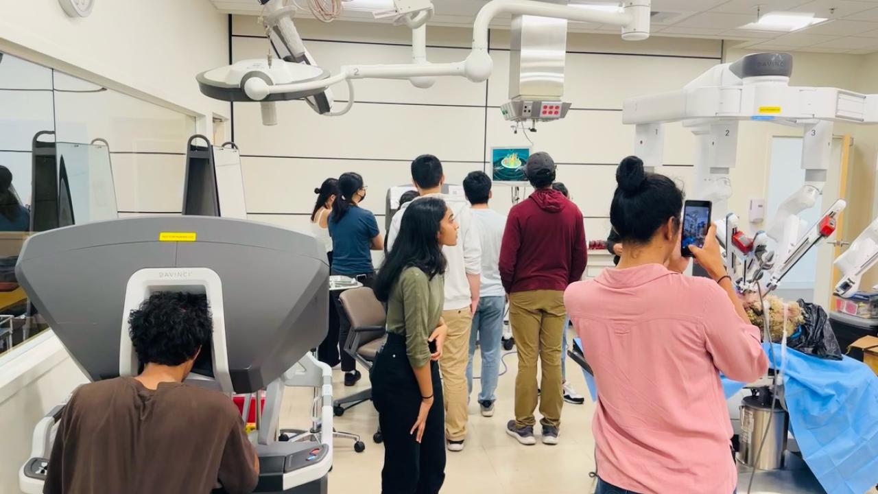 A group of people in a medical or research lab interacting with robotic surgical equipment. One person in the foreground is seated at a Da Vinci surgical console, while another is taking a photo with a smartphone. Several individuals in the background are gathered around a screen displaying a 3D model, while others are observing robotic arms covered in blue surgical drapes. The room is well-lit, with advanced medical technology and monitors mounted on the ceiling,