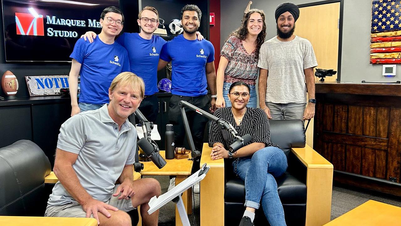 Mark Haney sits in a radio studio surrounded by several Square Solutions founders, students and entrepreneurs Henry Yu, Dillon Hill, Akshaj Raghavi, Sophie Bloyd, Harjn Bains and Deesha Patel. 