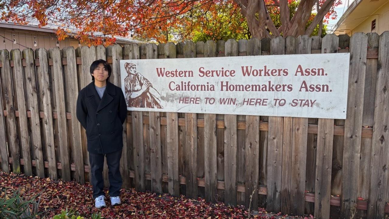 A photo of Seito Ezawa standing next to a fence with a poster of the Western Service Workers Association on a fall day with leaves on the ground.