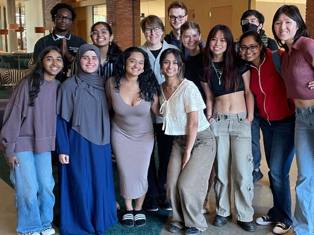 A group of 13 smiling students pose together for a photo in a lobby area of Aggie Square.