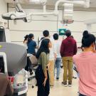 A group of people in a medical or research lab interacting with robotic surgical equipment. One person in the foreground is seated at a Da Vinci surgical console, while another is taking a photo with a smartphone. Several individuals in the background are gathered around a screen displaying a 3D model, while others are observing robotic arms covered in blue surgical drapes. The room is well-lit, with advanced medical technology and monitors mounted on the ceiling,