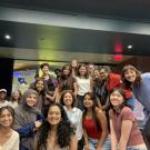 A large group of smiling young adults pose together indoors in a well-lit lounge or common area with dark ceilings and modern decor. The group consists of approximately twenty people, some seated and others standing closely behind them. They appear to be part of a class or program, with several individuals leaning in cheerfully toward the camera. The atmosphere is friendly and relaxed, and the space behind them includes glass walls, soft lighting, and a mix of seating areas.